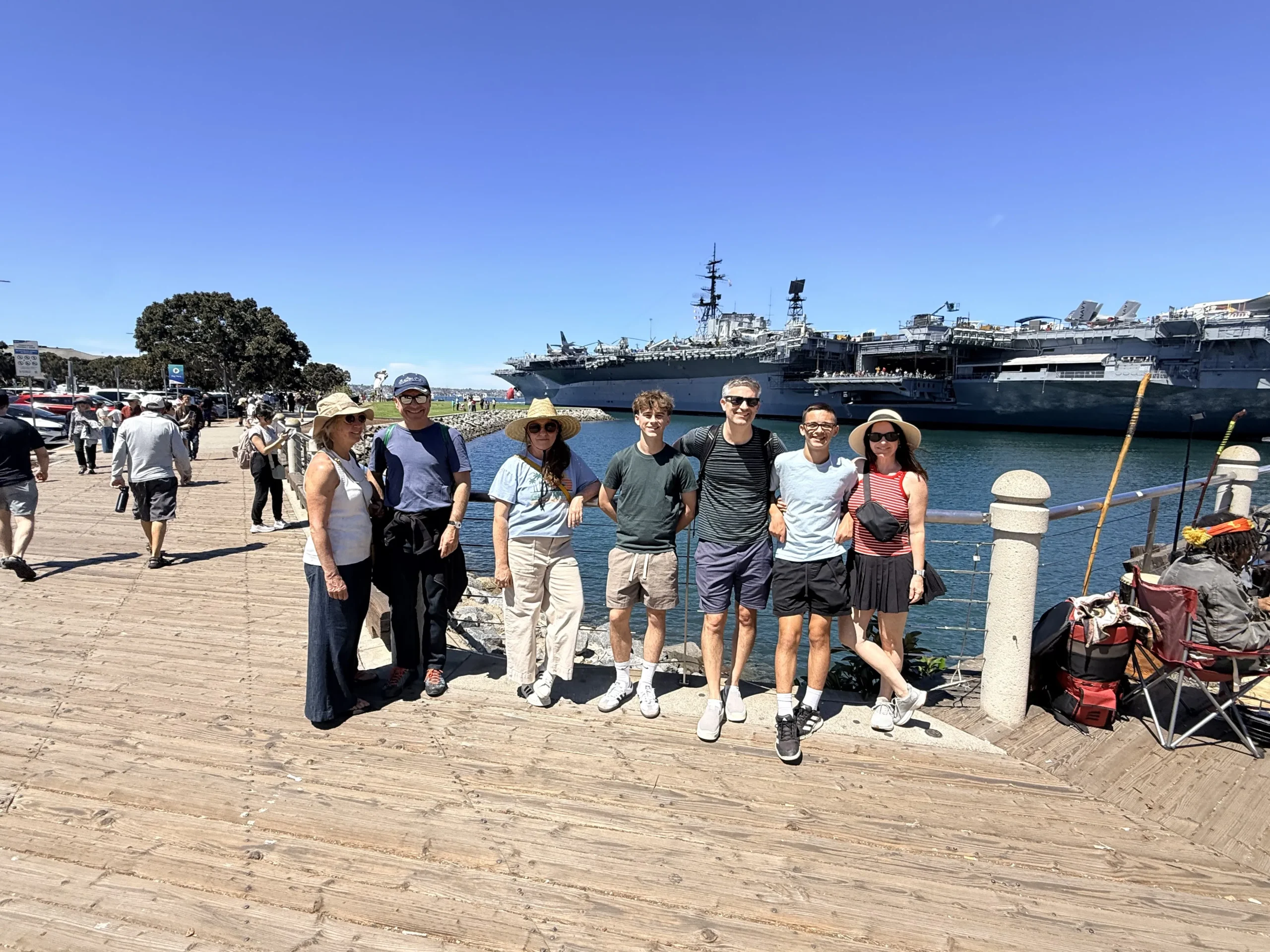 Six people on a sunny wooden pier with a massive aircraft carrier behind them.